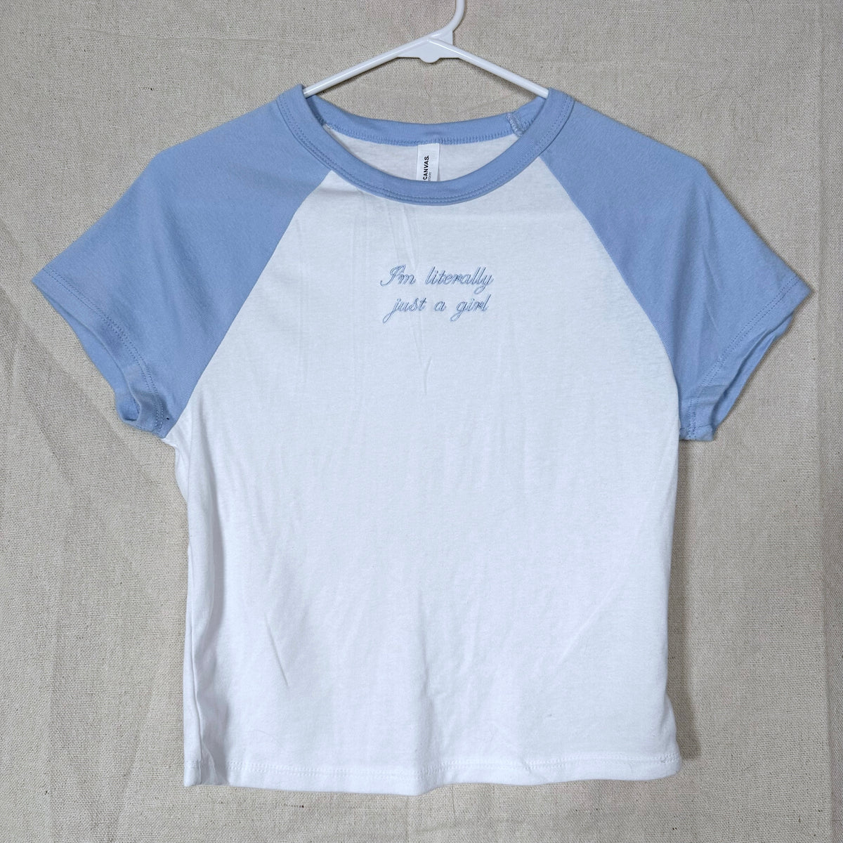 A raglan-style short-sleeve t-shirt hanging on a white hanger against a beige fabric backdrop. The shirt has a white body and light blue sleeves, with small blue cursive text on the chest reading 'I'm literally just a girl'.