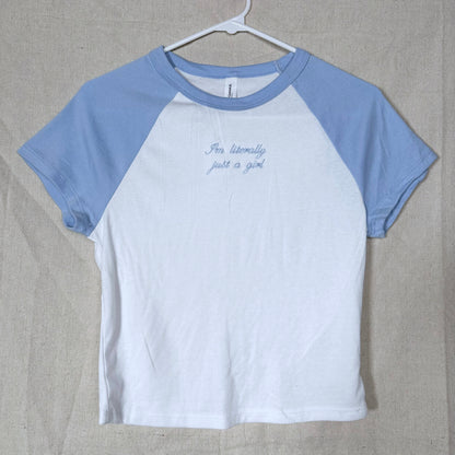 A raglan-style short-sleeve t-shirt hanging on a white hanger against a beige fabric backdrop. The shirt has a white body and light blue sleeves, with small blue cursive text on the chest reading 'I'm literally just a girl'.